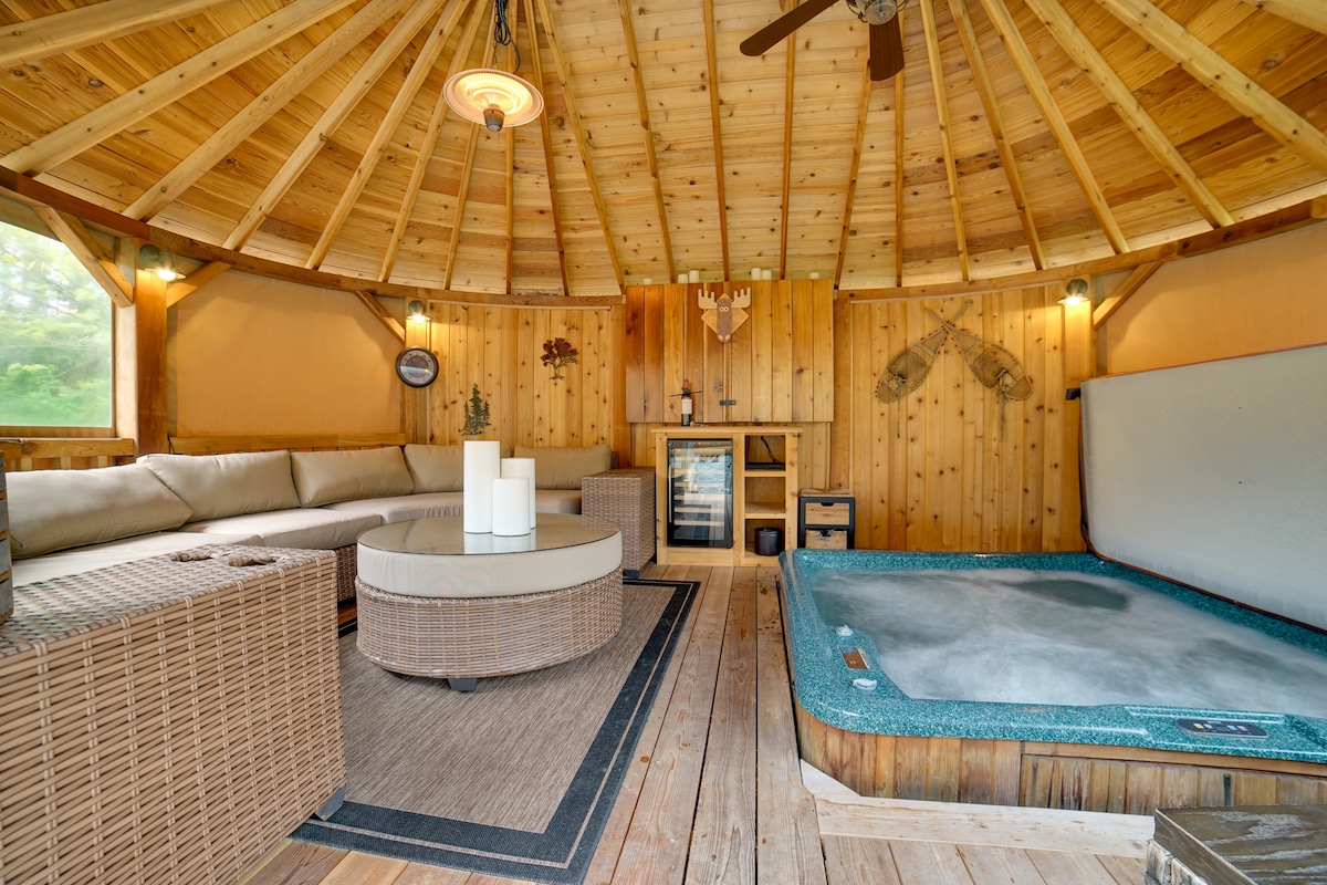 The gazebo features a circular layout with wooden beams overhead, complemented by a spacious seating area with plush cushions. A coffee table sits in the center, alongside a private hot tub. A beverage fridge and decor accentuate the rustic charm of the space.