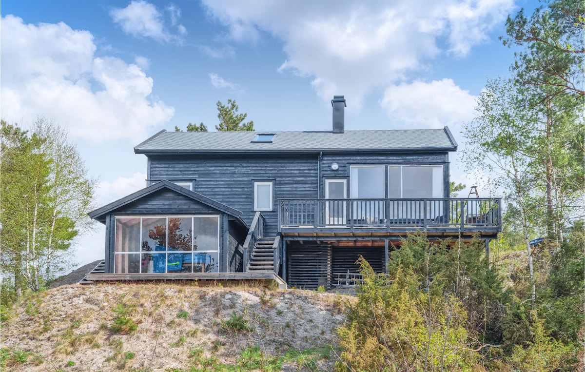 A charming wooden house is situated on a hill, featuring a large deck accessible from the main entrance. Expansive glass windows allow for ample natural light, highlighting the serene surroundings of trees and the landscape.