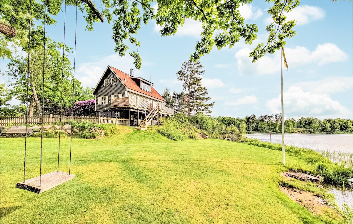 A charming house stands near Lake Hjälmsjön, surrounded by lush greenery. A wooden swing hangs from a tree in the foreground, while the house features a spacious deck with a railing, offering scenic views of the lake and landscape.