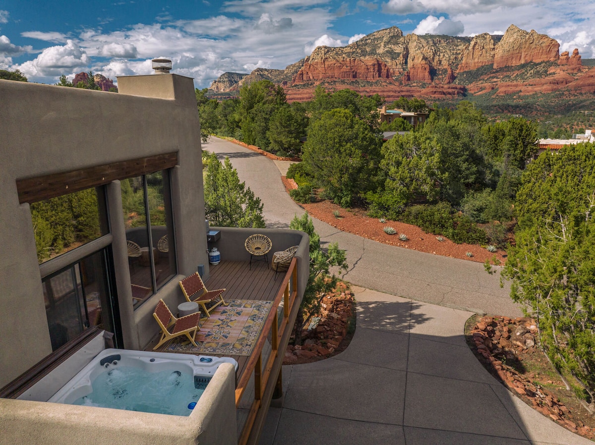 An outdoor deck features a private hot tub surrounded by scenic views of red rock formations and greenery. Two cozy lounge chairs rest nearby, facing the landscaped pathway that leads into the serene landscape.