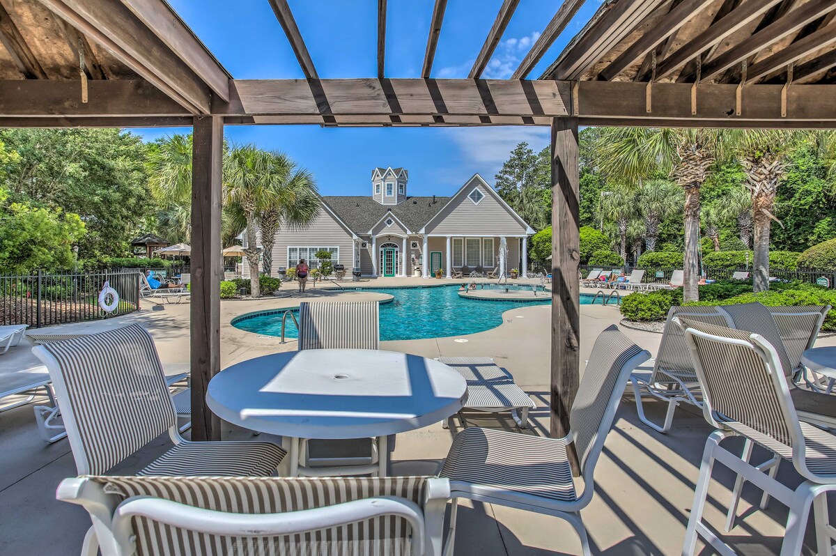 A shaded outdoor area features a round table surrounded by several white lounge chairs. The inviting pool is visible in the background, framed by palm trees and lush greenery. A clubhouse can be seen beyond the pool, contributing to a relaxing atmosphere.