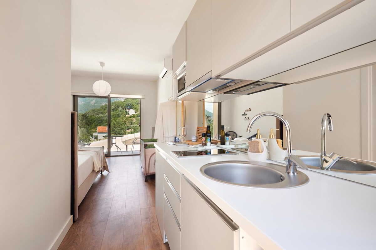 A modern kitchenette features a double sink and sleek cabinetry, leading into a light-filled sleeping area with a spacious bed. Large glass doors provide access to a private balcony with views of the surrounding greenery, enhancing the overall open layout.