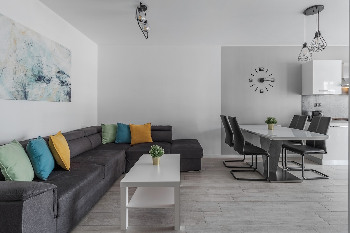 A modern living area features a spacious dark sofa adorned with colorful cushions. A minimalistic coffee table sits in front, complemented by a dining space with stylish chairs. The wall clock adds a touch of elegance, while natural light enhances the room's inviting atmosphere.