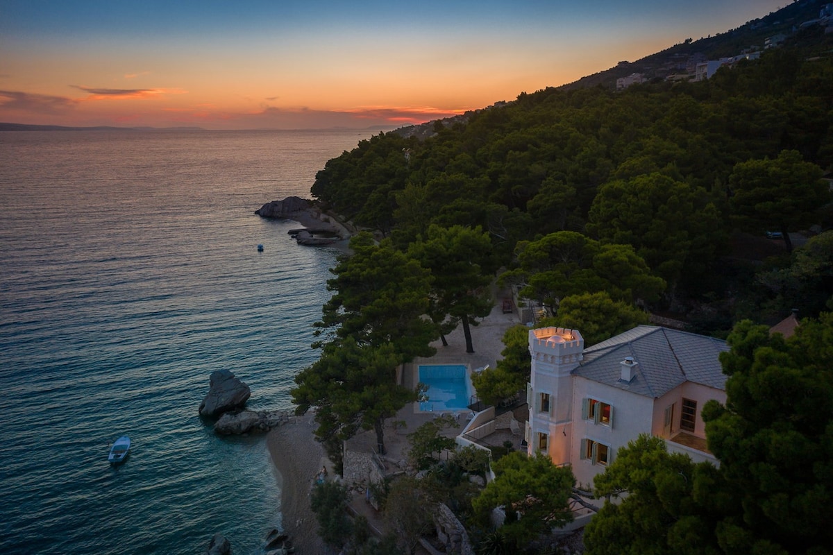 An aerial view captures the Luxury Villa Sea Palace, nestled among pine trees by the shimmering Adriatic Sea. The outdoor swimming pool glistens under a vibrant sunset, while the villa's unique tower stands out against the serene coastal landscape.