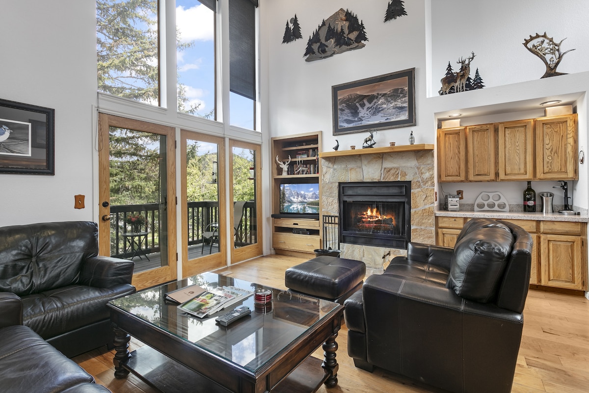 A spacious living area features a cozy fireplace surrounded by natural stone. Large windows allow natural light to illuminate the room, offering views of the outdoors. Comfortable leather seating is arranged around a coffee table, with a kitchen visible in the background.