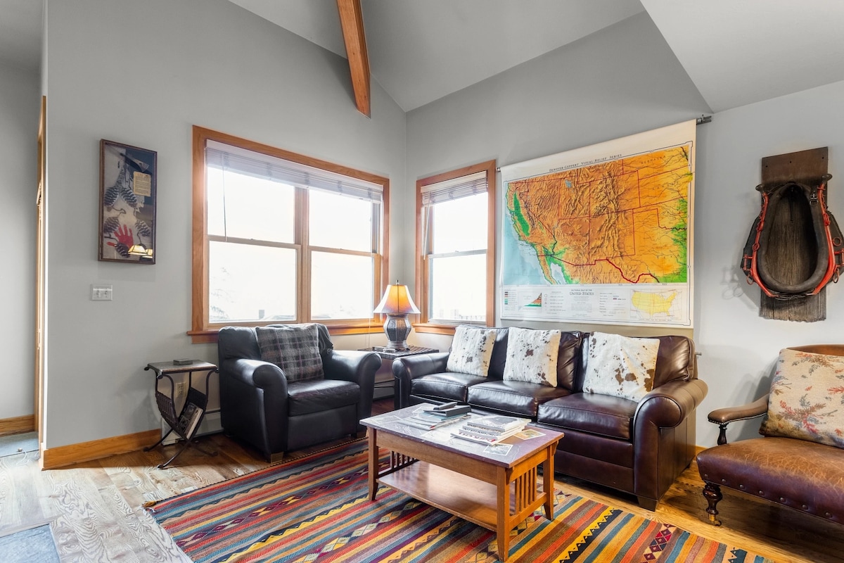 A living area features comfortable seating with a dark leather sofa and armchairs. A wooden coffee table holds materials for leisure activities. Two large windows allow natural light to fill the space, complemented by a colorful area rug and a detailed wall map.