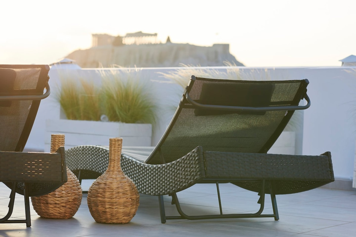 Two loungers are positioned on a private roof garden, surrounded by potted grass. The soft sunlight casts a warm glow on the space, highlighting woven baskets placed nearby, creating a serene setting perfect for relaxation.
