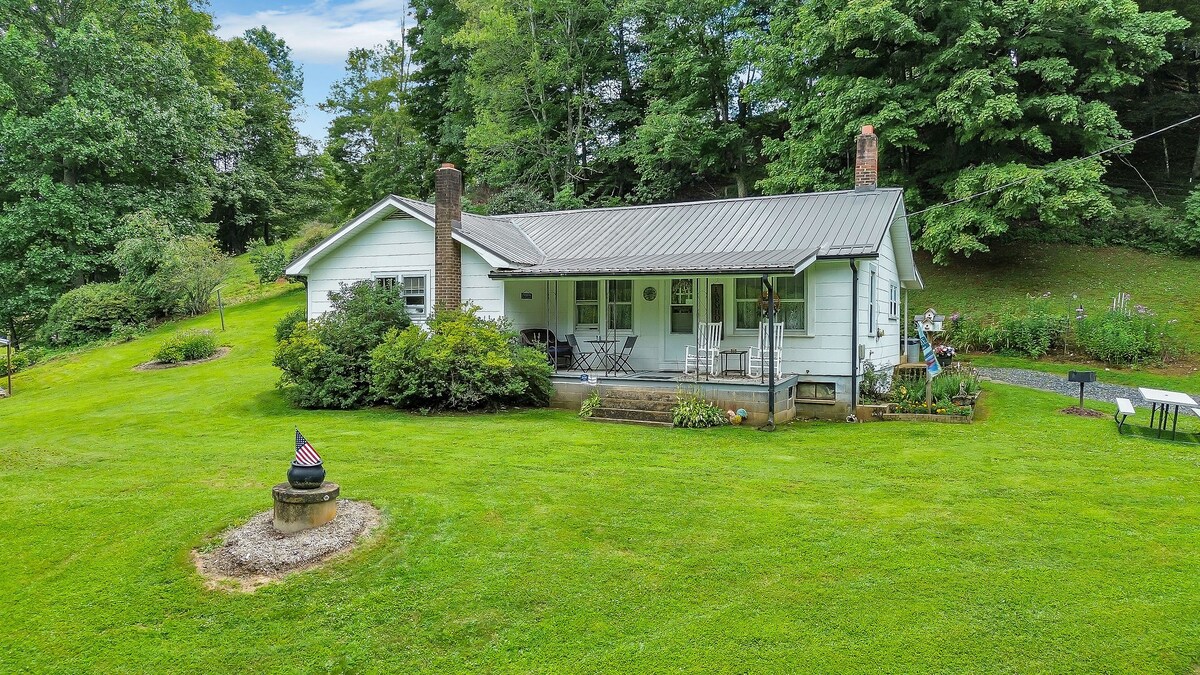 The vintage farmhouse is set on a spacious green lawn surrounded by trees. A welcoming front porch is lined with chairs, providing a view of the garden. A fire pit with an American flag stands in the yard, complemented by flower beds and a table placed nearby.