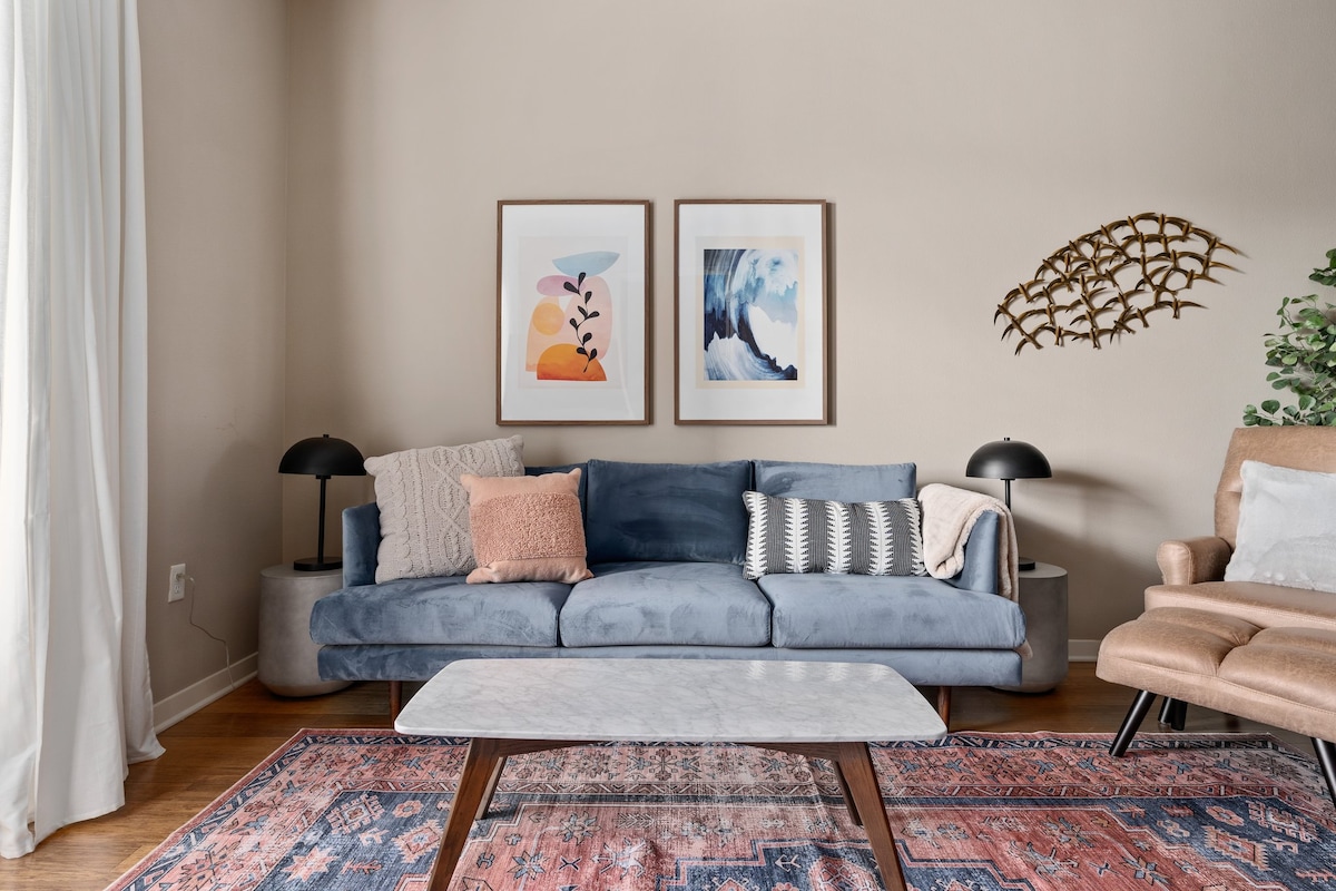 A inviting living area features a comfortable blue sofa with decorative cushions, flanked by stylish lamps. Above, two framed artworks are displayed on the beige wall, while a light wood coffee table sits atop a colorful area rug, enhancing the ambiance.