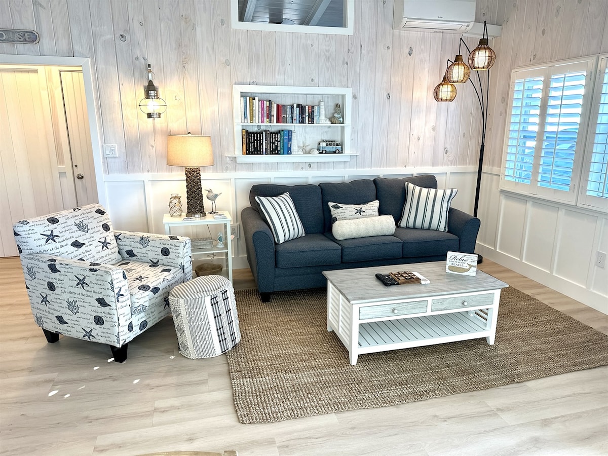 A light-filled living room features a comfortable navy sofa with decorative cushions, a patterned armchair, and a round accent table. A coffee table rests on a woven rug, while built-in shelves display books. Soft lighting is provided by stylish pendant fixtures and table lamps.