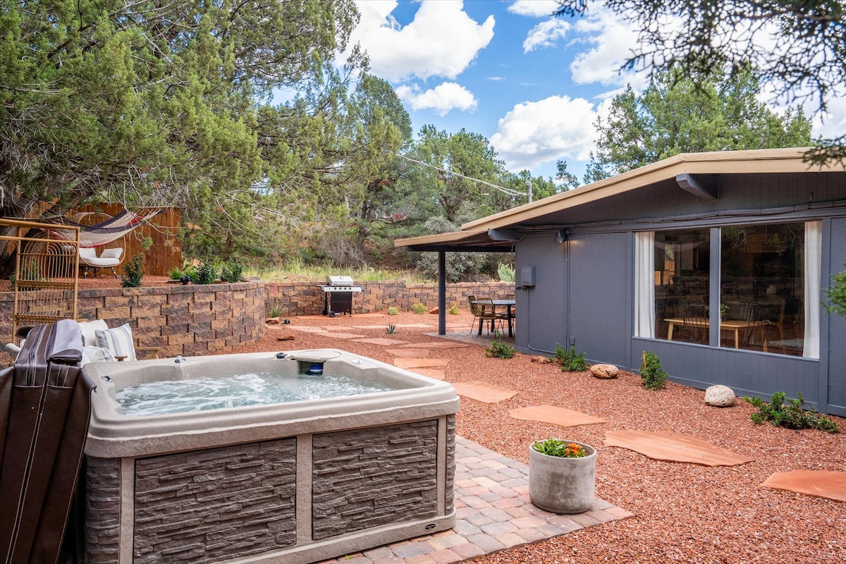 A private backyard features a hot tub surrounded by natural landscaping, terracotta pavers, and decorative stones. The hot tub is positioned next to a seating area with a dining table. In the background, a grill is visible near the entrance of the home.