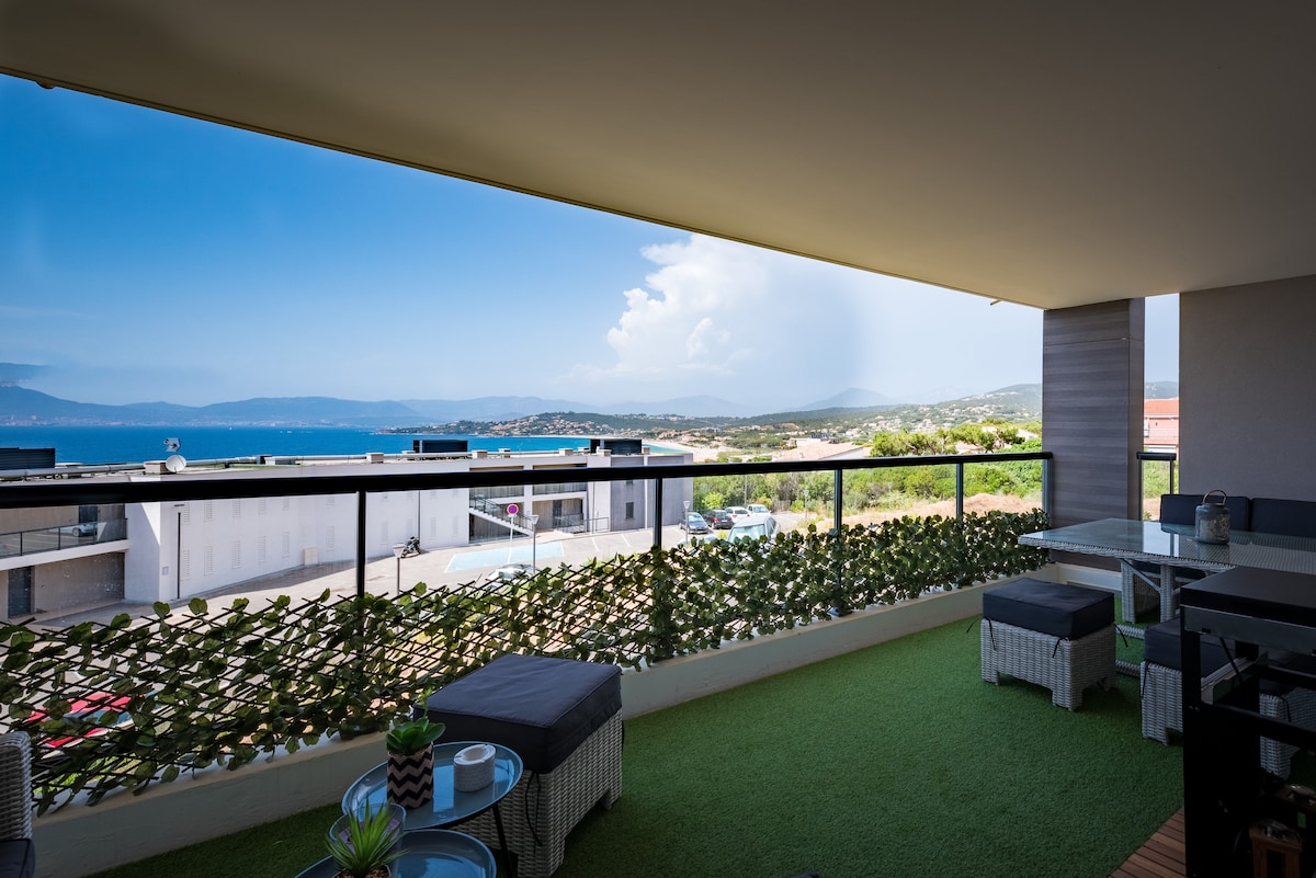 A spacious balcony offers a serene view of the sea and distant mountains. Furnished with comfortable seating and a dining area, the outdoor space is enhanced by greenery along the railing. Natural light floods the area, creating an inviting atmosphere for relaxation.