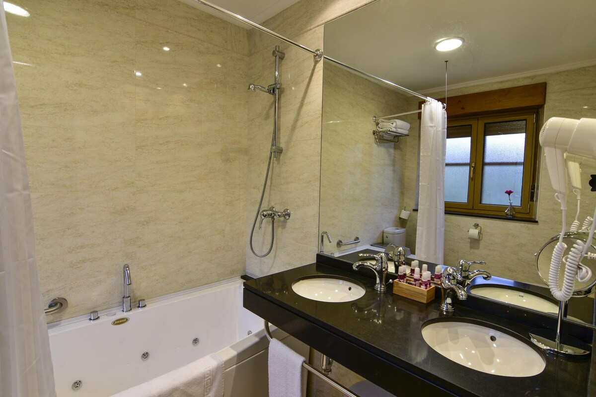 A well-appointed bathroom features a spacious bathtub alongside a double vanity with two sinks. A large mirror reflects natural light from the window, while toiletries are neatly arranged on a wooden tray. A hairdryer and towel racks are also present.