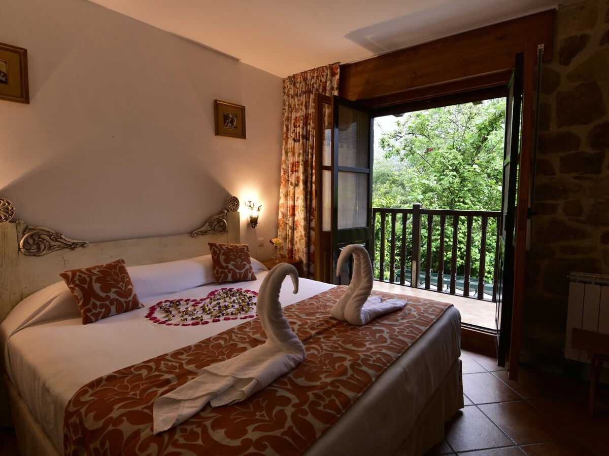 A spacious bed is centered in a softly lit room, adorned with decorative pillows. Towels are arranged in a swan shape on the bed. A balcony door opens to views of lush greenery, bringing natural light into the inviting space.