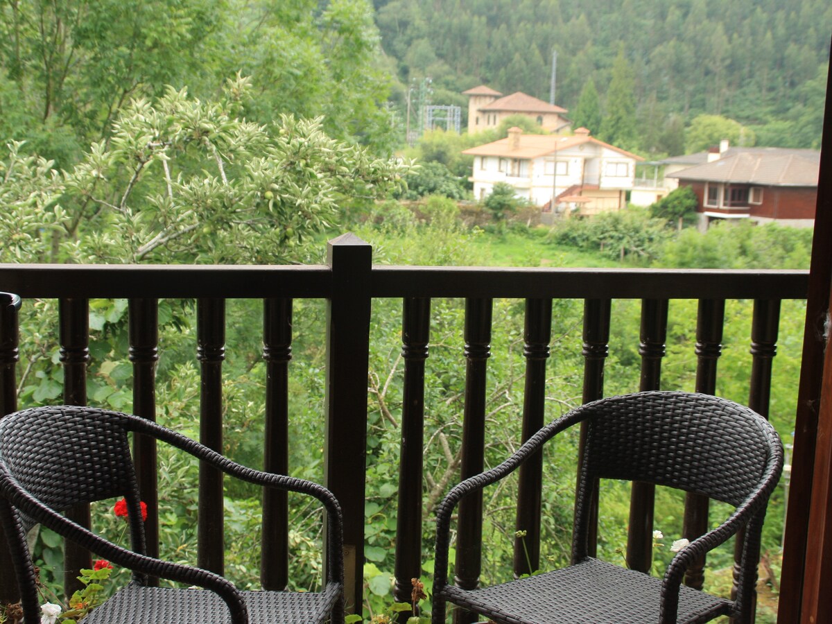 A balcony railing made of dark wood encloses two black wicker chairs, providing a relaxing space to enjoy views of the surrounding greenery and mountains. Lush trees and distant houses create a tranquil rural atmosphere.