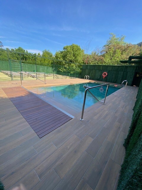 A clear swimming pool is enclosed by a wooden deck and surrounded by greenery. Safety railings are visible alongside the pool, and a lifebuoy hangs on the wall. The bright sky reflects onto the water, creating a calm ambiance.