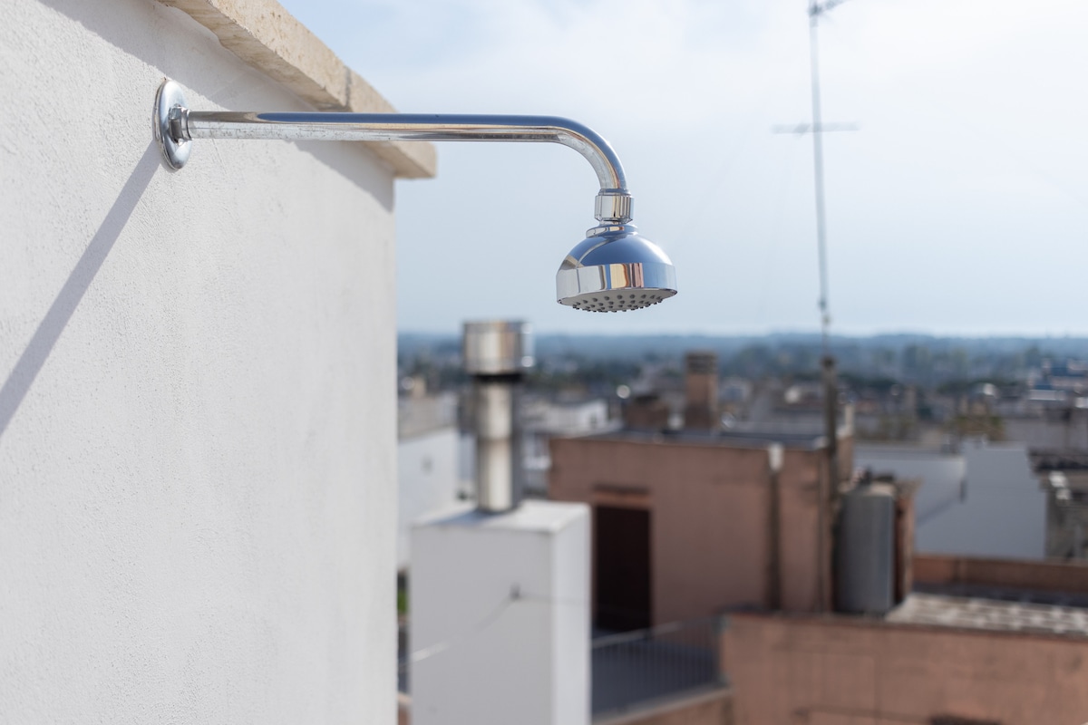 An outdoor shower fixture is installed on a white wall, featuring a sleek, metallic showerhead. The background showcases a view of surrounding rooftops and distant hills, under a clear sky.
