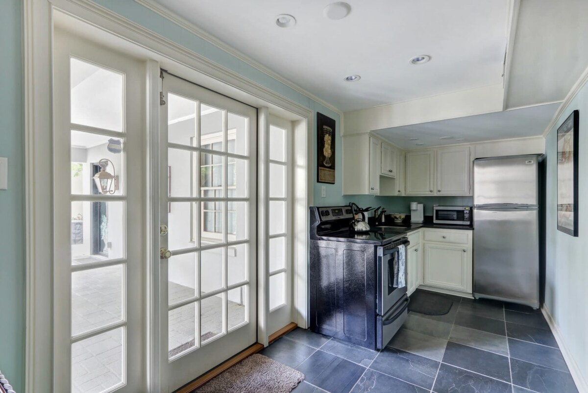 The modernized kitchen features a black stove, stainless-steel refrigerator, and granite countertops. French doors invite natural light into the space, providing a view of the enclosed garden patio. Light-colored cabinetry complements the sleek, dark flooring.