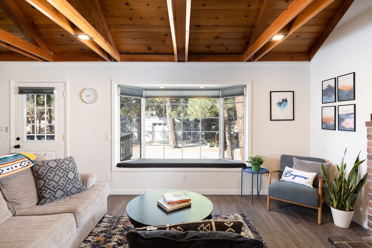 A living area features an open beam ceiling and large windows allowing natural light. A cozy couch invites relaxation, accompanied by a blue armchair. A decorative rug covers the floor, while a small table holds magazines. Potted greenery adds a touch of nature to the space.