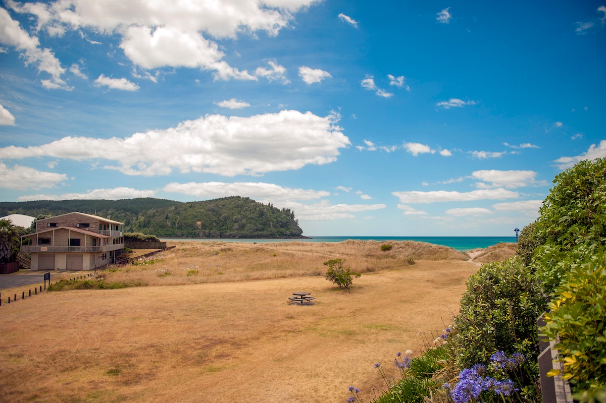 A wide, open field stretches toward the coastline, marked by sparse vegetation. The ocean glimmers under a bright blue sky with a few scattered clouds. In the distance, a gentle hill rises, with a structure partially visible to the left, suggesting a peaceful natural setting.