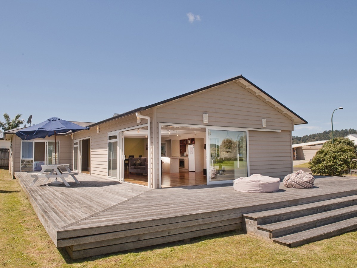 The exterior view of the holiday home reveals a modern structure with large glass doors leading to a spacious deck. Outdoor seating arrangements, including a picnic table and lounge chairs, are visible. The deck extends over a grassy area, providing a relaxing outdoor space.