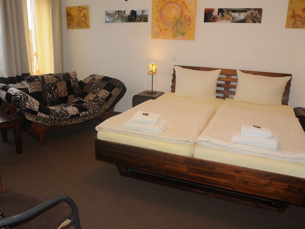 A spacious room is furnished with a large double bed and a comfortable sleeping couch. A wooden lamp adds subtle lighting, while artwork decorates the walls, enhancing the aesthetic. Soft, neutral tones are complemented by natural light flowing through the curtains.