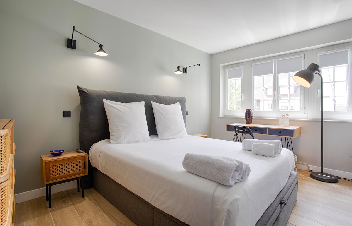 A bright bedroom features a spacious bed with two white pillows and neatly rolled towels. Wall sconces provide ambient lighting, while a desk and chair are positioned by large windows, allowing for natural light to fill the room.