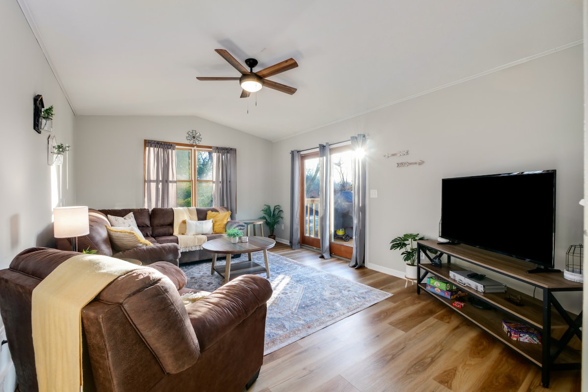 The living room features a comfortable seating area with a large sectional sofa and a coffee table. Natural light floods the space through large windows, and a ceiling fan adds to the ambiance. Double doors lead to an outdoor deck, enhancing the connection to nature.