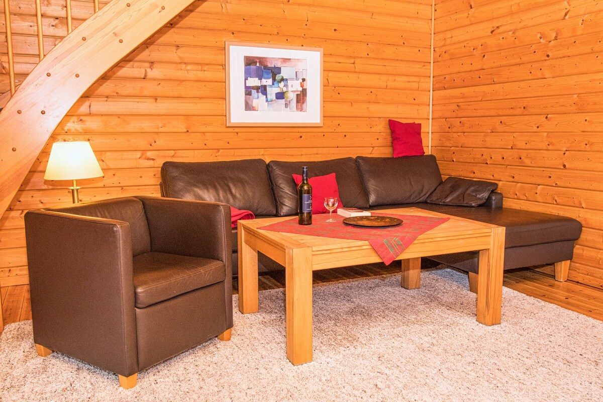 A cozy living area features warm natural wood walls and soft lighting. A sectional sofa, complemented by a comfortable armchair, faces a low wooden coffee table. A decorative piece rests on the table, and a framed artwork adds a splash of color to the space.