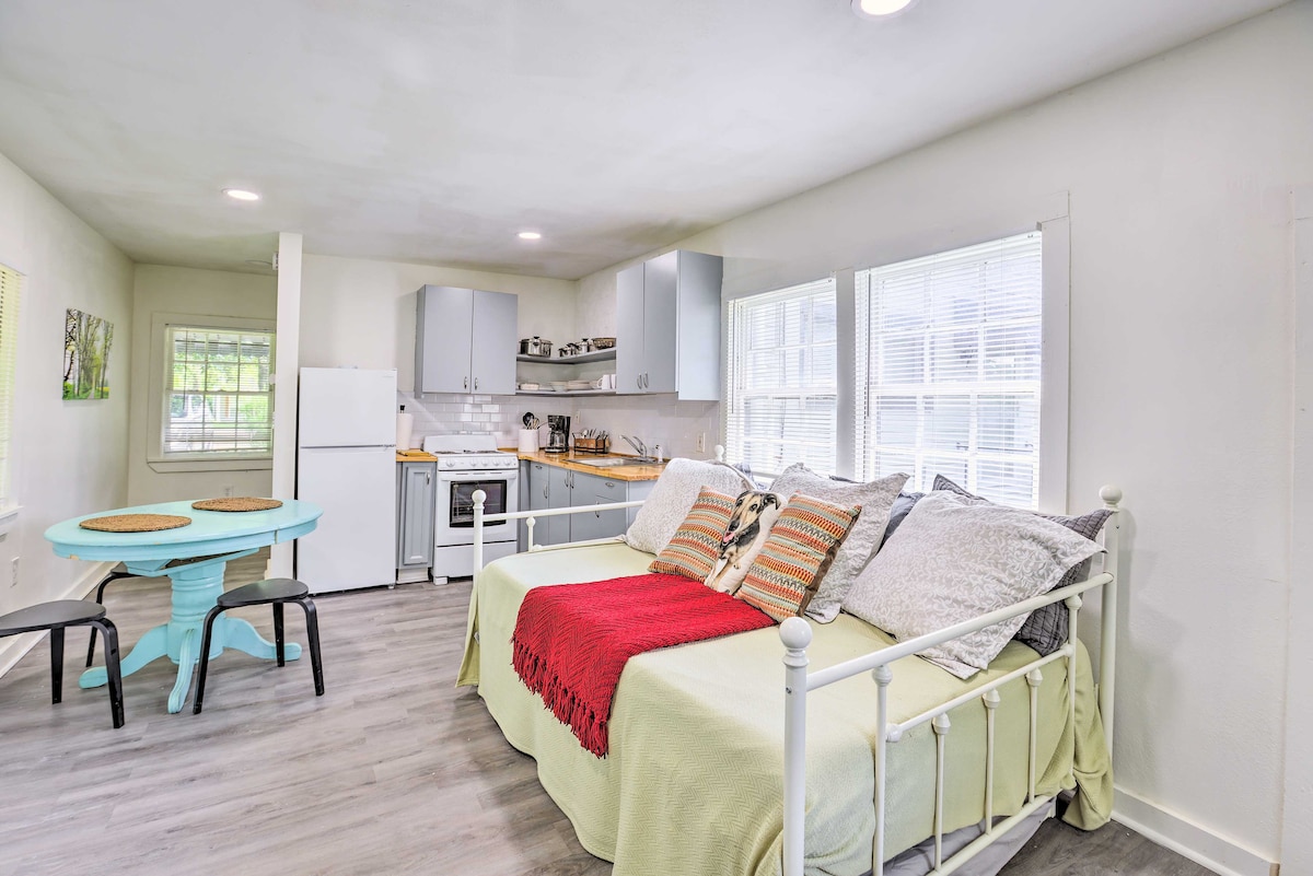 A light-filled studio space features a full daybed with a cozy twin trundle underneath, adorned with decorative pillows. A small round dining table with two chairs is visible, alongside a kitchen equipped with modern appliances and ample counter space. Windows allow for natural light.