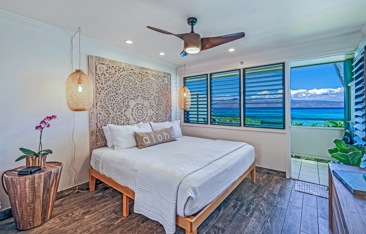 A spacious studio features a king-sized bed with neutral bedding, complemented by an artistic wall hanging. Large windows provide views of the ocean, and natural light fills the room. A fan and two light fixtures enhance the peaceful ambiance, while a small side table adds functionality.