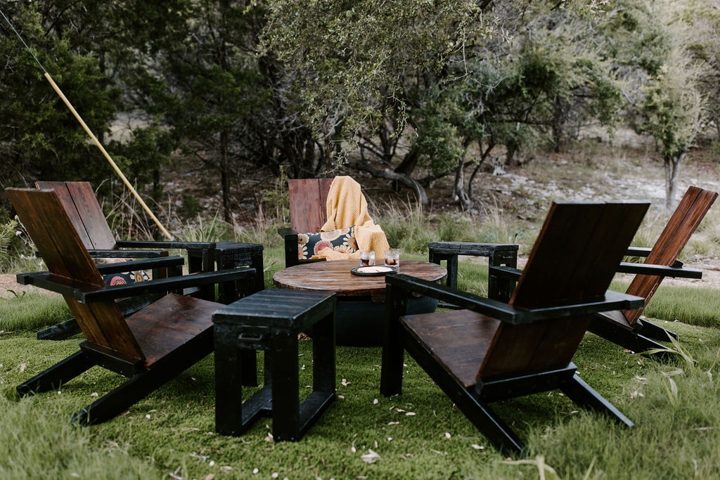 A rustic outdoor seating area is showcased, featuring four wooden chairs arranged around a circular table. A blanket is draped over one chair, creating a cozy touch. The setting is surrounded by lush greenery, enhancing the natural ambiance.