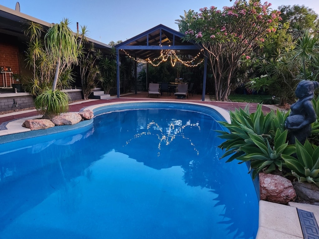 A serene swimming pool is framed by lush plants and a covered patio area, featuring comfortable seating. Soft lighting drapes around the structure, creating a welcoming ambiance. The clear blue water reflects the surrounding greenery and evening sky.