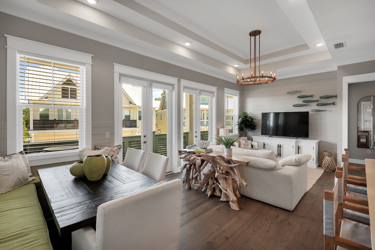 An open-concept living area displays a mix of natural materials and modern design. Large windows allow ample light, while a cozy sofa and unique coffee table invite relaxation. A wooden dining table accommodates family meals, and a TV sits centrally on a minimalist console.