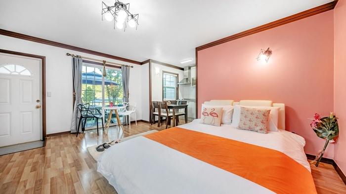 A spacious sleeping area features a large bed with an orange throw, complemented by colorful decorative cushions. Natural light enters through the window, illuminating the light wood floor. A small dining area with a table and chairs is visible in the background.