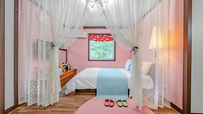 A soft pink bedroom features a spacious bed draped with light curtains. A bedside table holds decorative items, while a lamp emits gentle light. A circular rug in muted tones is positioned on the wooden floor, and colorful slippers are neatly placed nearby.