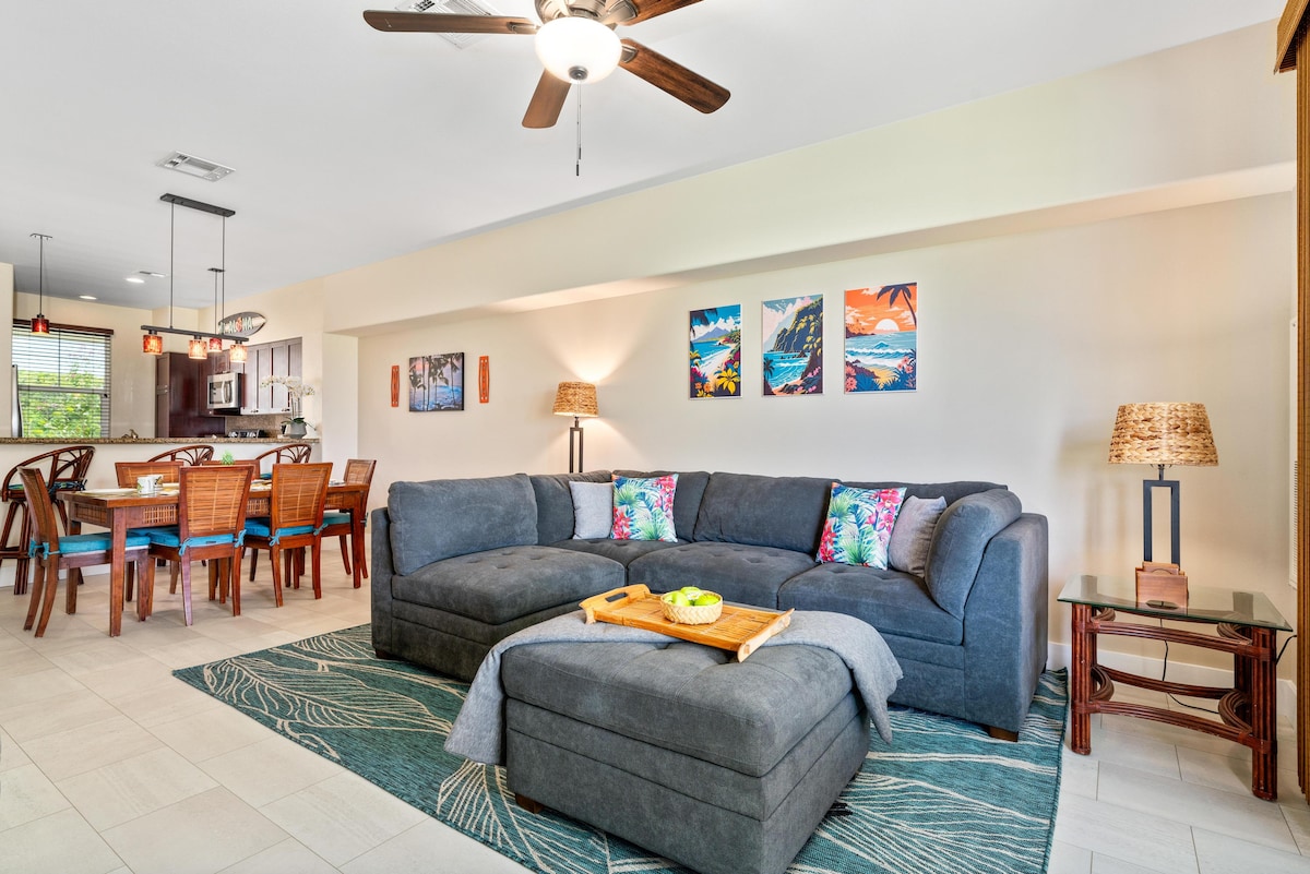 A spacious living area is highlighted by a large sectional sofa adorned with decorative pillows. A coffee table sits nearby, topped with fruit. The walls feature colorful artwork, while a dining area with wooden chairs is visible in the background, creating a harmonious space for gatherings.