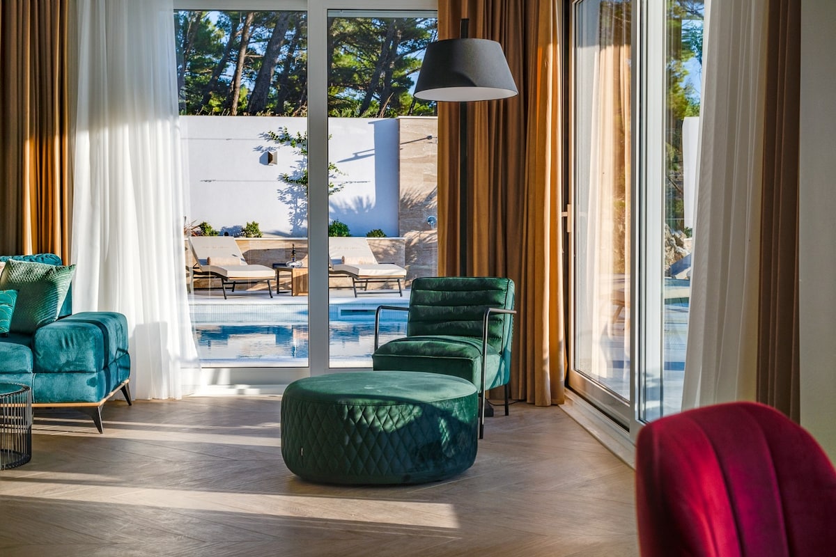 A spacious living area features large glass doors that open to the pool area, allowing natural light to enhance the interior. Comfortable seating includes a green ottoman and stylish armchairs, while sheer curtains softly frame the picturesque outdoor view.