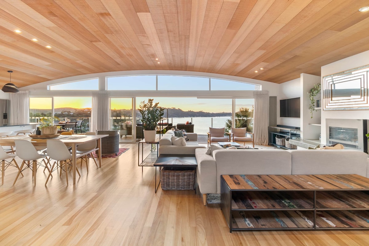 An open-plan living area features warm wooden ceilings and expansive floor-to-ceiling windows, showcasing stunning sunset views over the water. A comfortable seating arrangement and a dining table are present, creating a welcoming space for relaxation and gatherings.