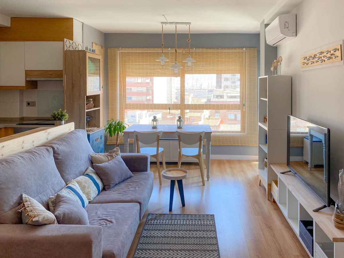 A well-lit living area features a comfortable sofa with decorative pillows and a coffee table. Natural light filters through the window covered with bamboo blinds. A dining table for four is visible, complemented by shelving that adds functional decor.