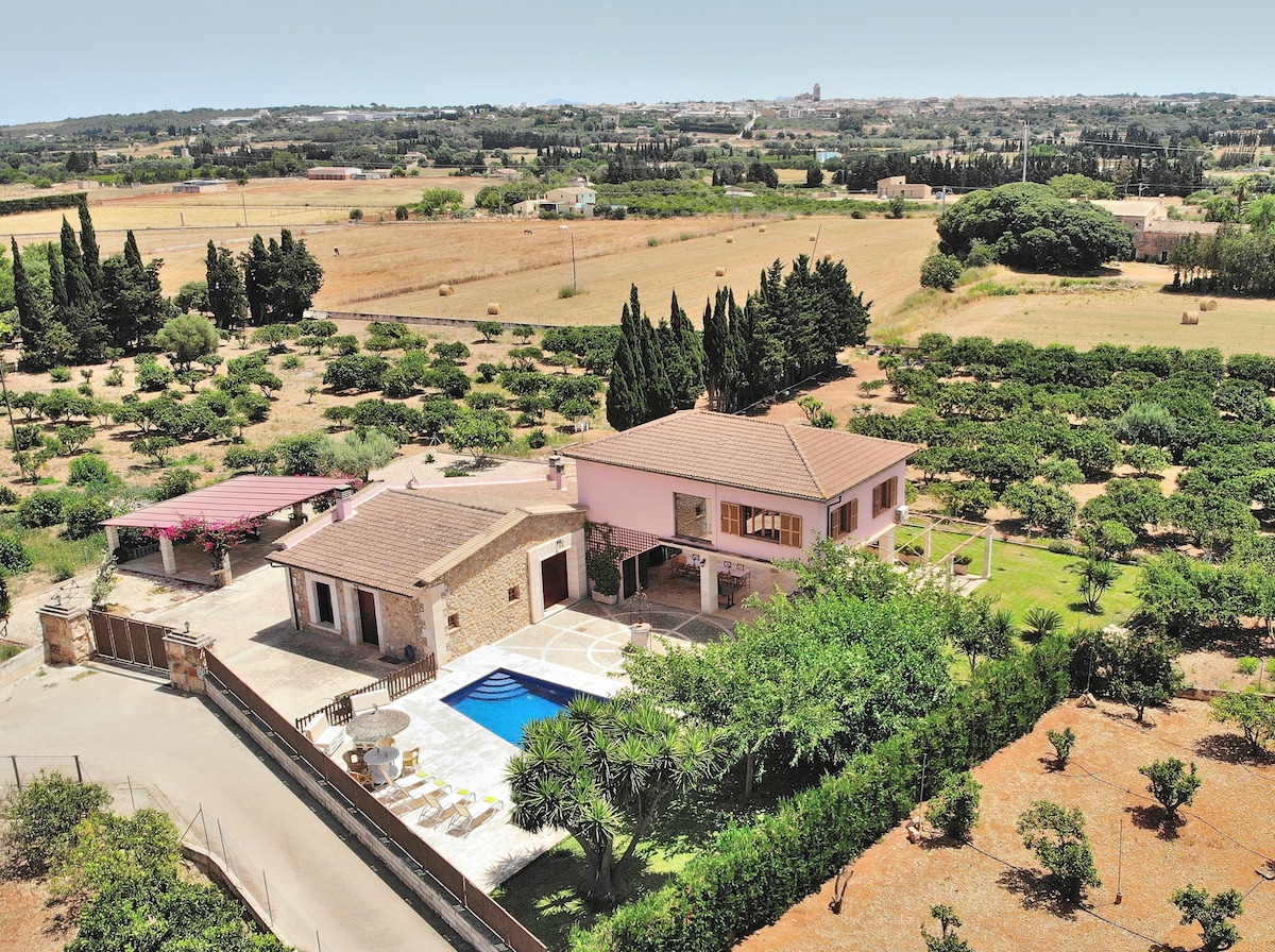 An aerial view captures the villa surrounded by lush greenery and open fields. The private swimming pool is centered beside the terrace, while the home displays a harmonious blend of stone and pastel architecture, framed by trees that enhance the peaceful rural setting.