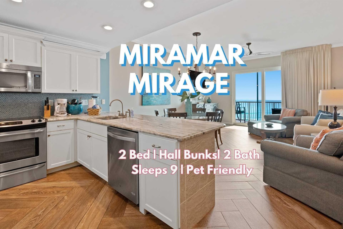 The modern kitchen features stainless steel appliances, including a stove and refrigerator, complemented by a spacious island with bar seating. An open living area presents comfortable seating options, with large glass doors offering a view of the Gulf and access to a private balcony.