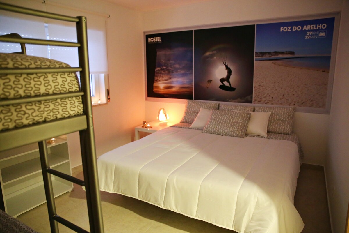 A well-lit room features a spacious bed with a simple white coverlet and decorative pillows. A metal bunk bed is positioned alongside, while a bedside table with a lamp adds a soft glow. The wall showcases scenic photographs, enhancing the room's serene atmosphere.