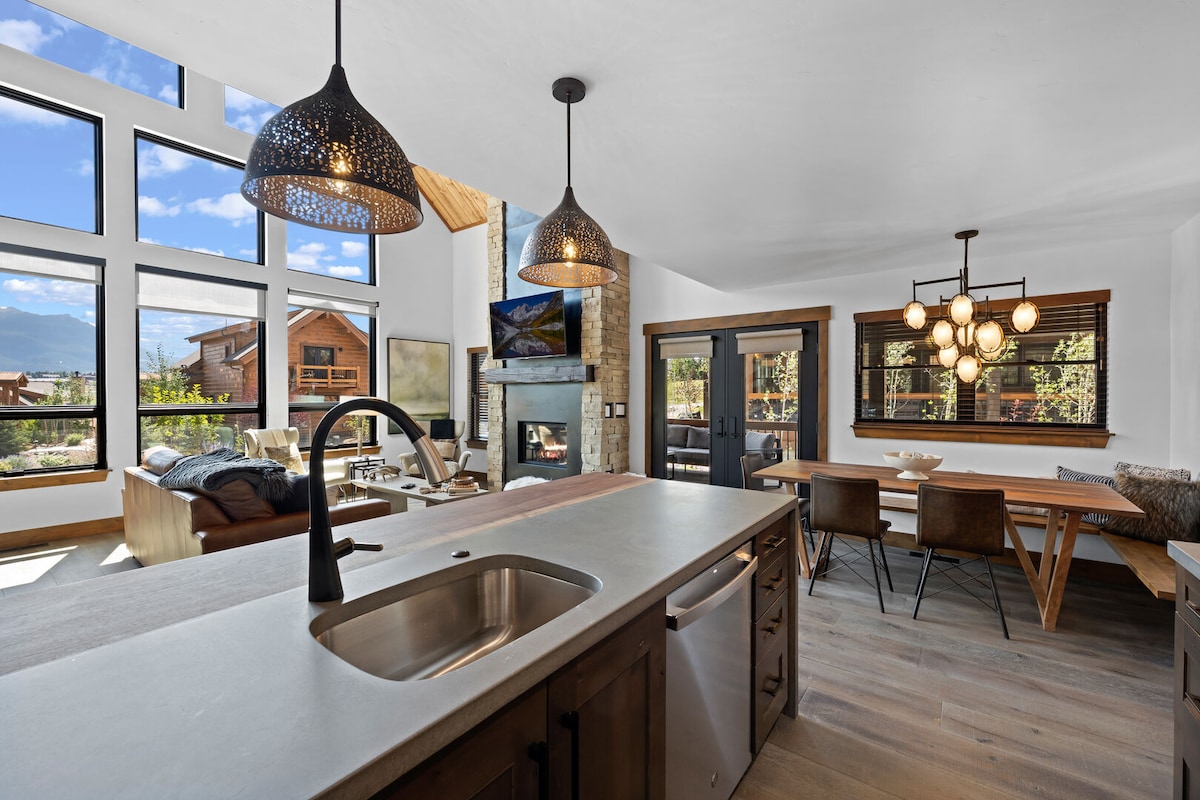 An open floorplan highlights a modern kitchen with sleek cabinets and a large island featuring a sink. Natural light fills the space through large windows, showcasing views of the mountains. A dining table for six and a cozy seating area with a stone fireplace create an inviting atmosphere.