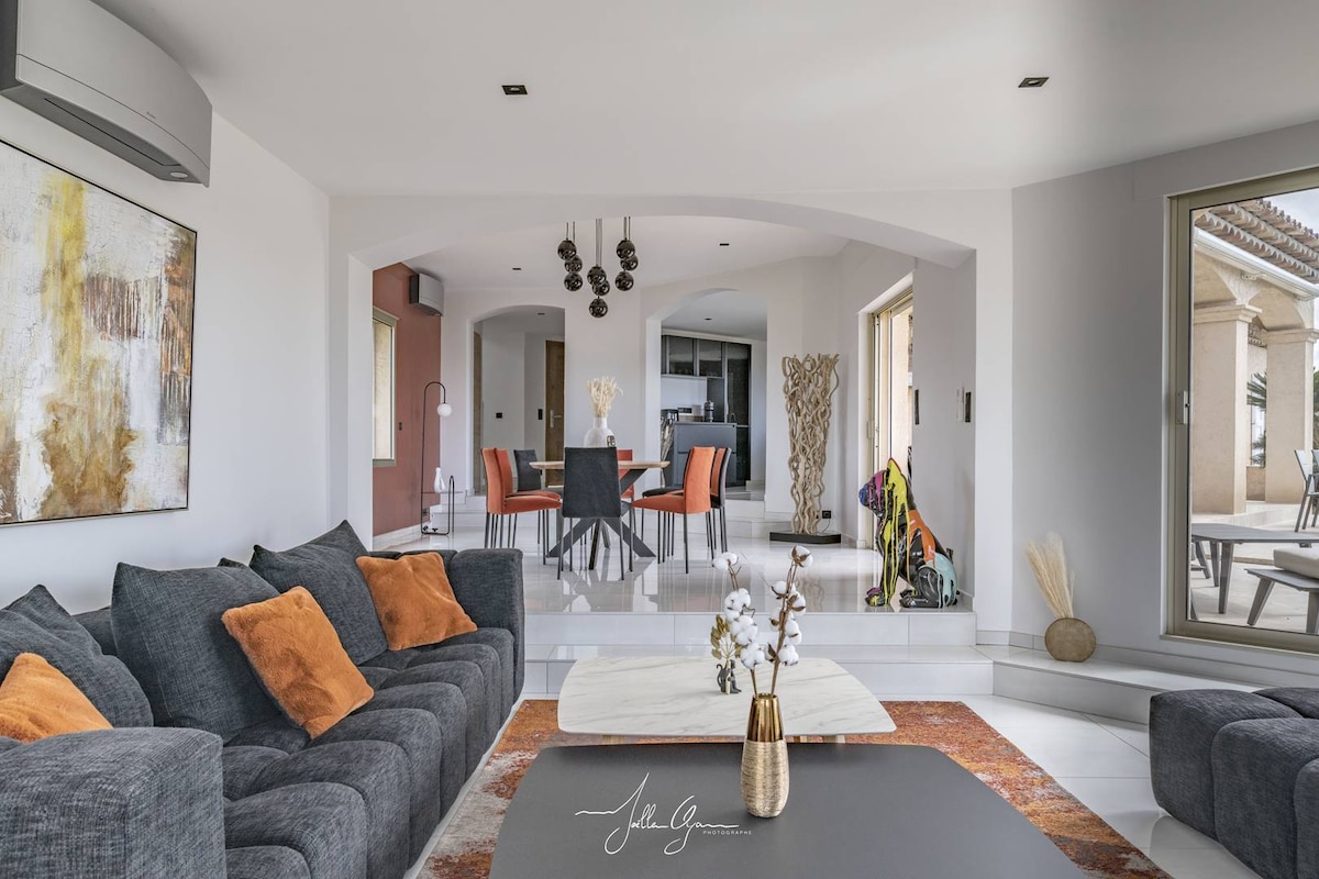 A spacious living room showcases a large, dark upholstered sofa with colorful accent pillows. Near the sofa, a minimalist coffee table is positioned on a stylish rug. Behind, a dining area is visible, featuring modern chairs and a round table, with large windows allowing natural light to flow in.