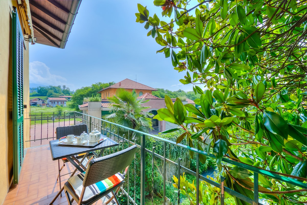 A small balcony is presented with a bistro table and chairs, surrounded by lush greenery. The view includes rooftops and trees, creating a serene atmosphere. The railing adds a layer of accessibility to this inviting outdoor space.