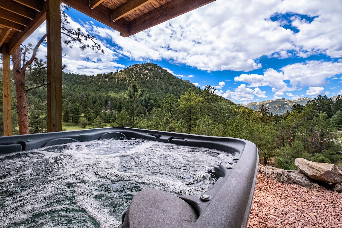 A hot tub is positioned on a wooden deck, offering a view of lush green mountains under a partly cloudy sky. Bubbles rise from the water, creating a serene atmosphere, while natural rock formations and trees frame the scene.