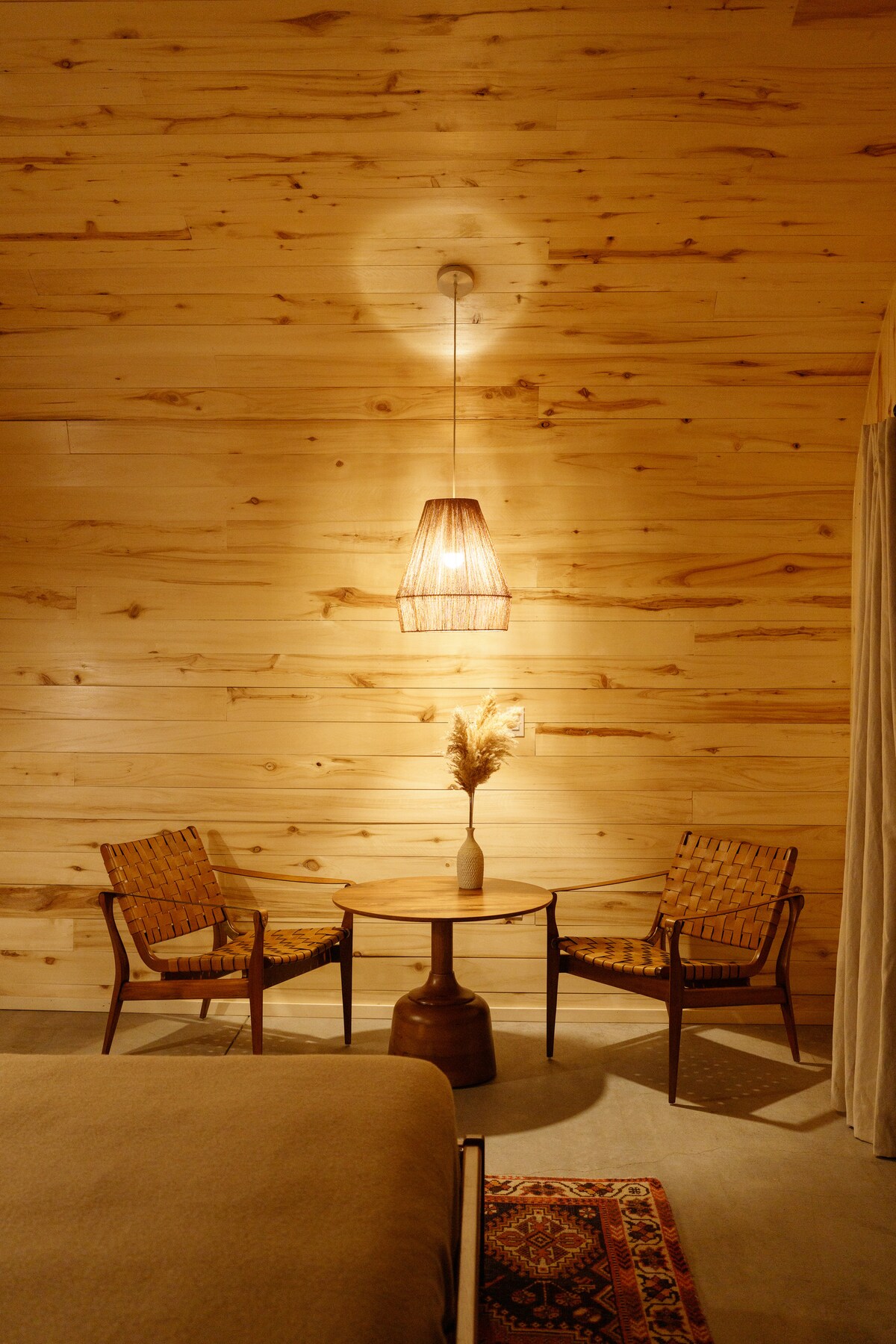 A serene corner of a room is highlighted by warm wooden walls. Two woven chairs flank a small table, adorned with a minimalist vase holding dried botanicals. A pendant light casts gentle illumination, creating a calm and inviting space.