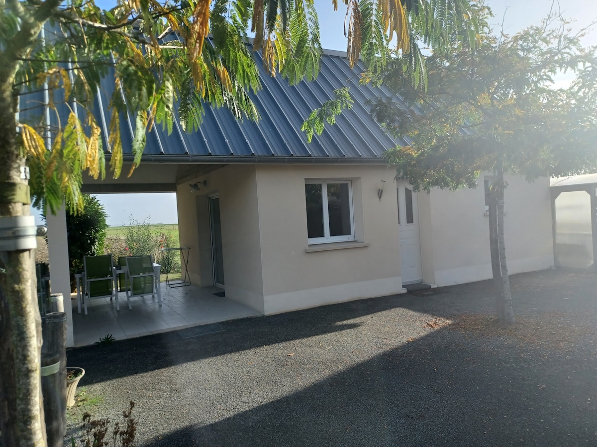 The exterior of a gite is viewed, showcasing a light-colored facade with a sloped metal roof. A covered terrace with dining furniture is seen to the left, while trees provide gentle shade in the surrounding area. The entrance door is visible, leading into the living space.