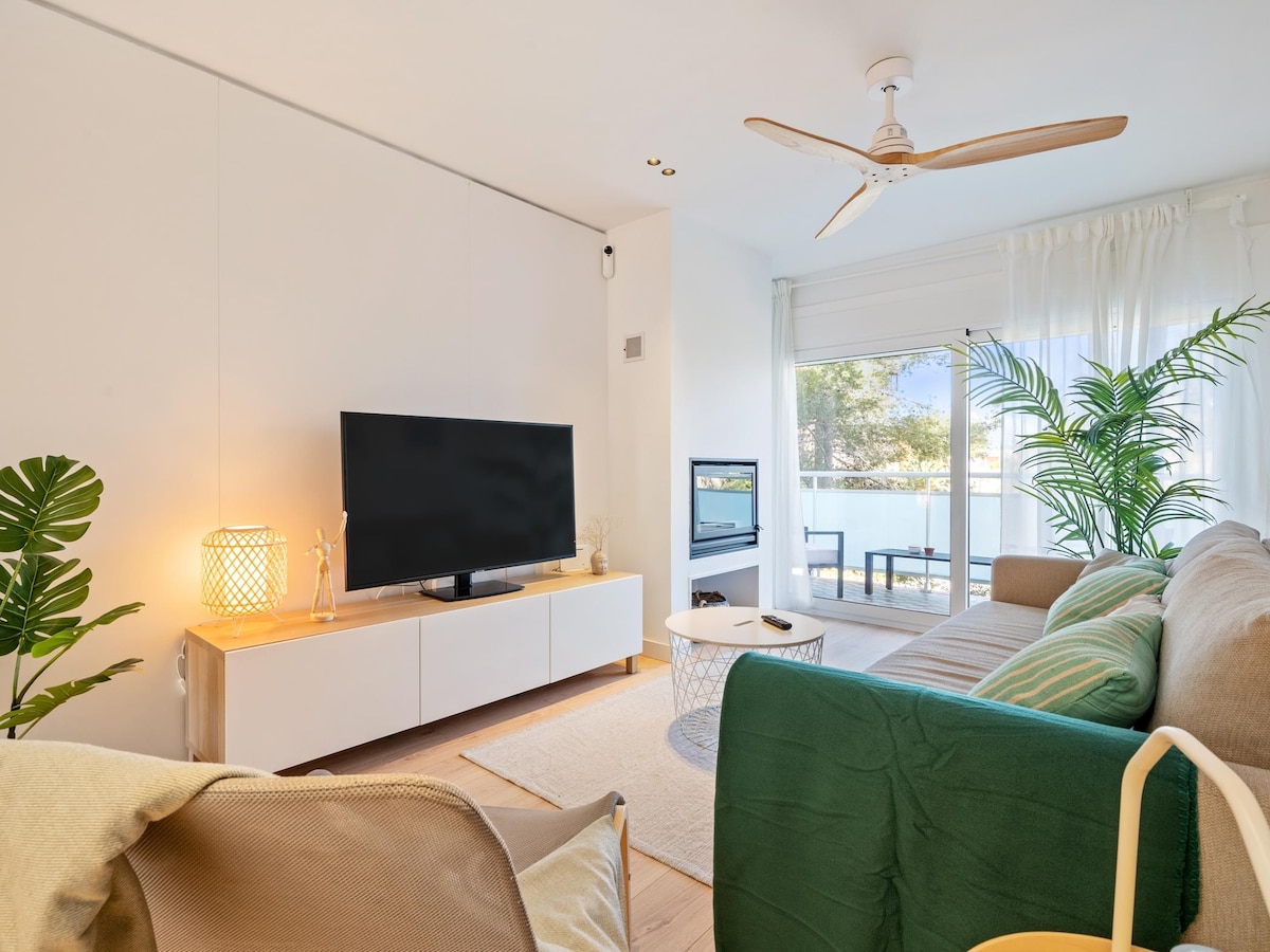 A cozy living room is showcased, featuring a large flat-screen TV positioned on a white cabinet. A comfortable sofa with decorative cushions and a light rug provides seating. A small coffee table sits in front, and a plant adds a touch of greenery. Natural light enters through the nearby window.